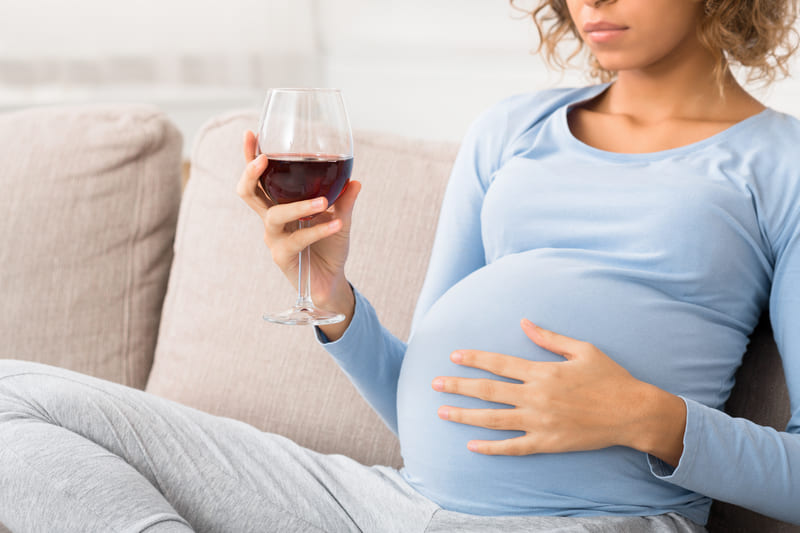 Kobieta w zaawansowanej ciąży siedzi na kanapie i trzyma lampkę czerwonego wina
